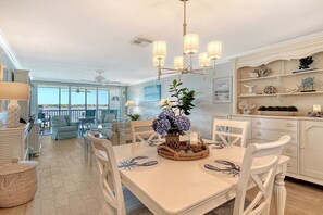 A dining area with a table set for four, surrounded by white chairs, a chandelier, and a view of the outdoors. - Sunrise Cove 308: Remodeled Stunning Coastal Waterfront Condo on Siesta Key (Sarasota)
