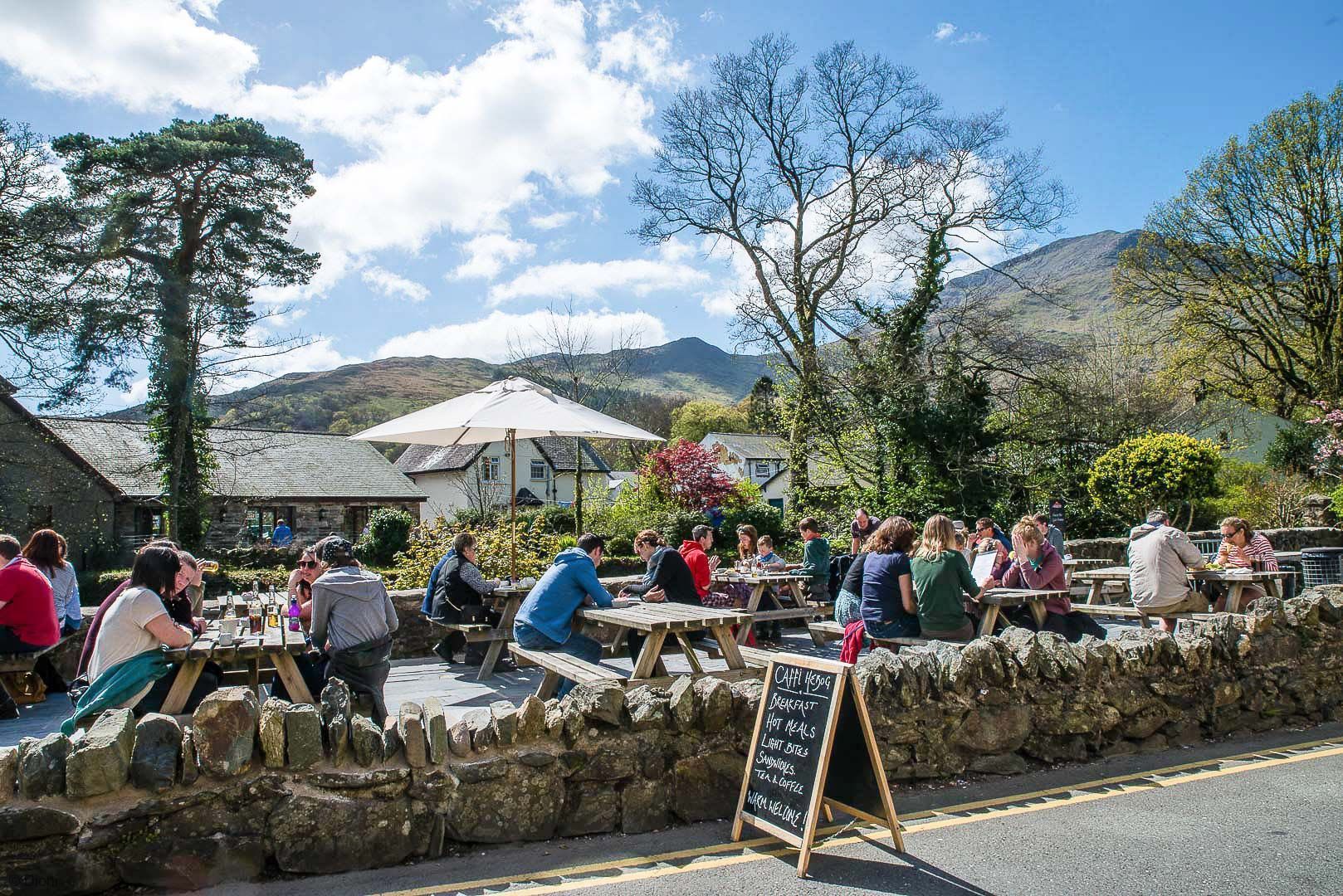 An outdoor dining area with stone walls and wooden tables, surrounded by trees and a mountainous backdrop.