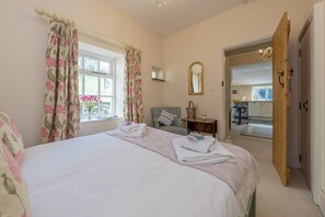 A bedroom with a bed, a bedside table, a chair, a mirror, and a view into the kitchen.