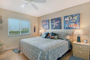 A bedroom with a bed, two bedside tables, a window with blinds, and artwork on the wall. - Sunrise Cove 402: Fully Remodeled Waterfront Condo on Siesta Key (Sarasota)