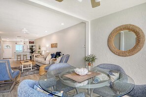Un salon moderne avec une table basse en verre, des fauteuils bleus, un canapé et une cuisine en arrière-plan.