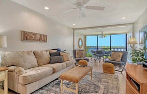 Un salon avec un canapé, un pouf et un ventilateur de plafond. Il y a un balcon avec vue.