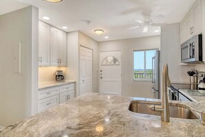 A kitchen with white cabinets, a marble countertop, a stainless steel sink, and a ceiling fan. - Sunrise Cove 403: Modern Waterfront Condo on Siesta Key with Murphy Bed (Sarasota)