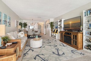 Ein Wohnzimmer mit einer Couch, einem Hocker und einem Fernseher auf einem Holzkasten. Auf dem Boden liegt ein Teppich mit Anker-Motiven.