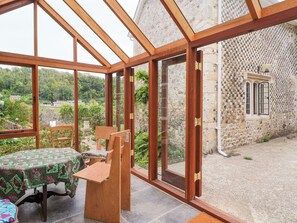 A conservatory with a table and chairs, a stone wall, and a view of the countryside. - Hole House (Seaton)