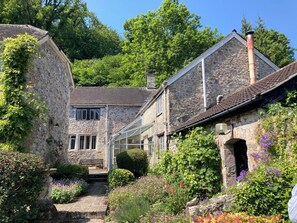 A stone building with a covered walkway, surrounded by lush greenery and flowering plants. - Hole House (Seaton)