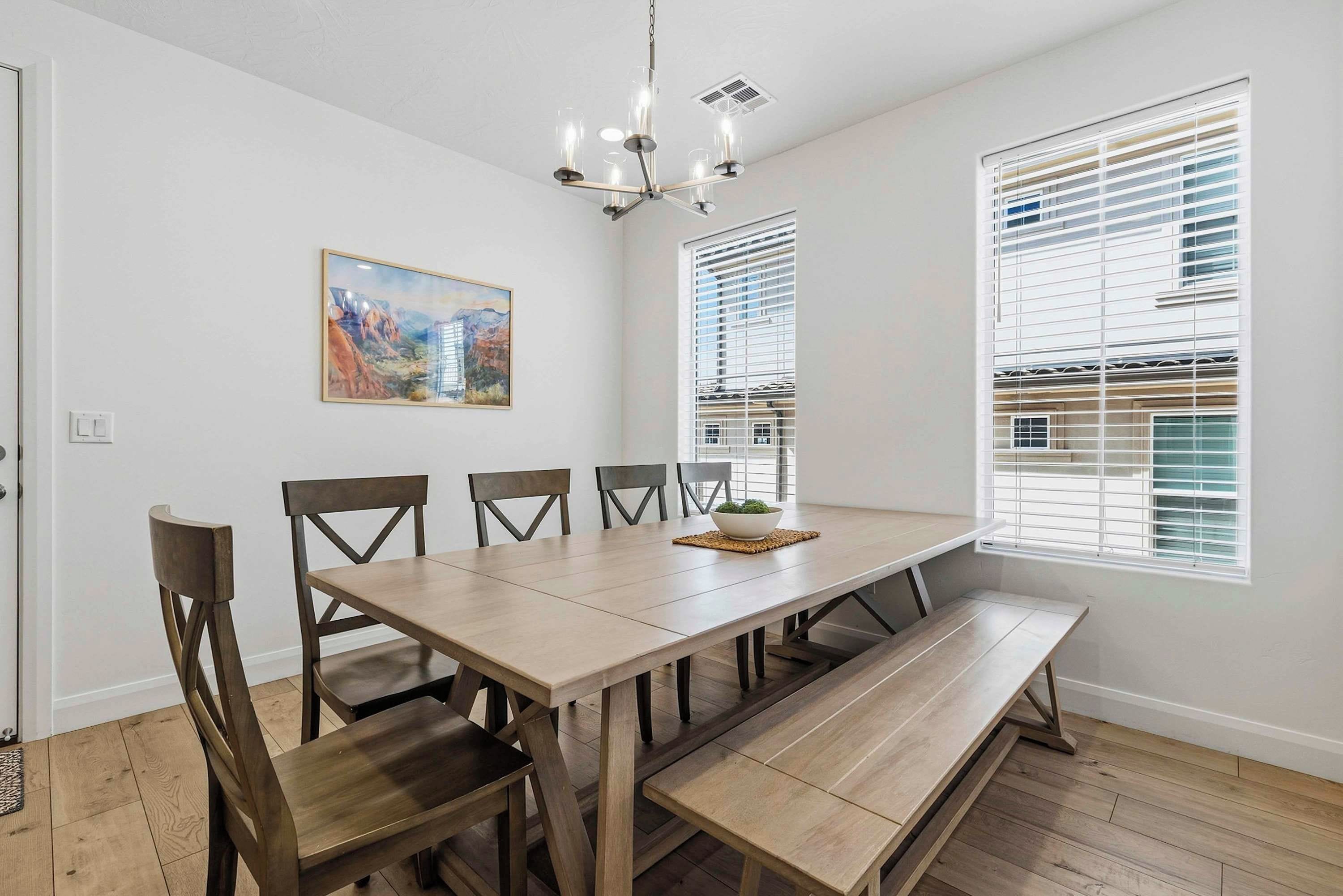 A dining area with a long wooden table, four chairs, a bench, a chandelier, and a window with blinds.