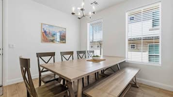 A dining area with a long wooden table, four chairs, a bench, a chandelier, and a window with blinds.