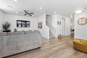 A modern living room with a grey sofa, a ceiling fan, a flat-screen TV, and a staircase leading to another room.