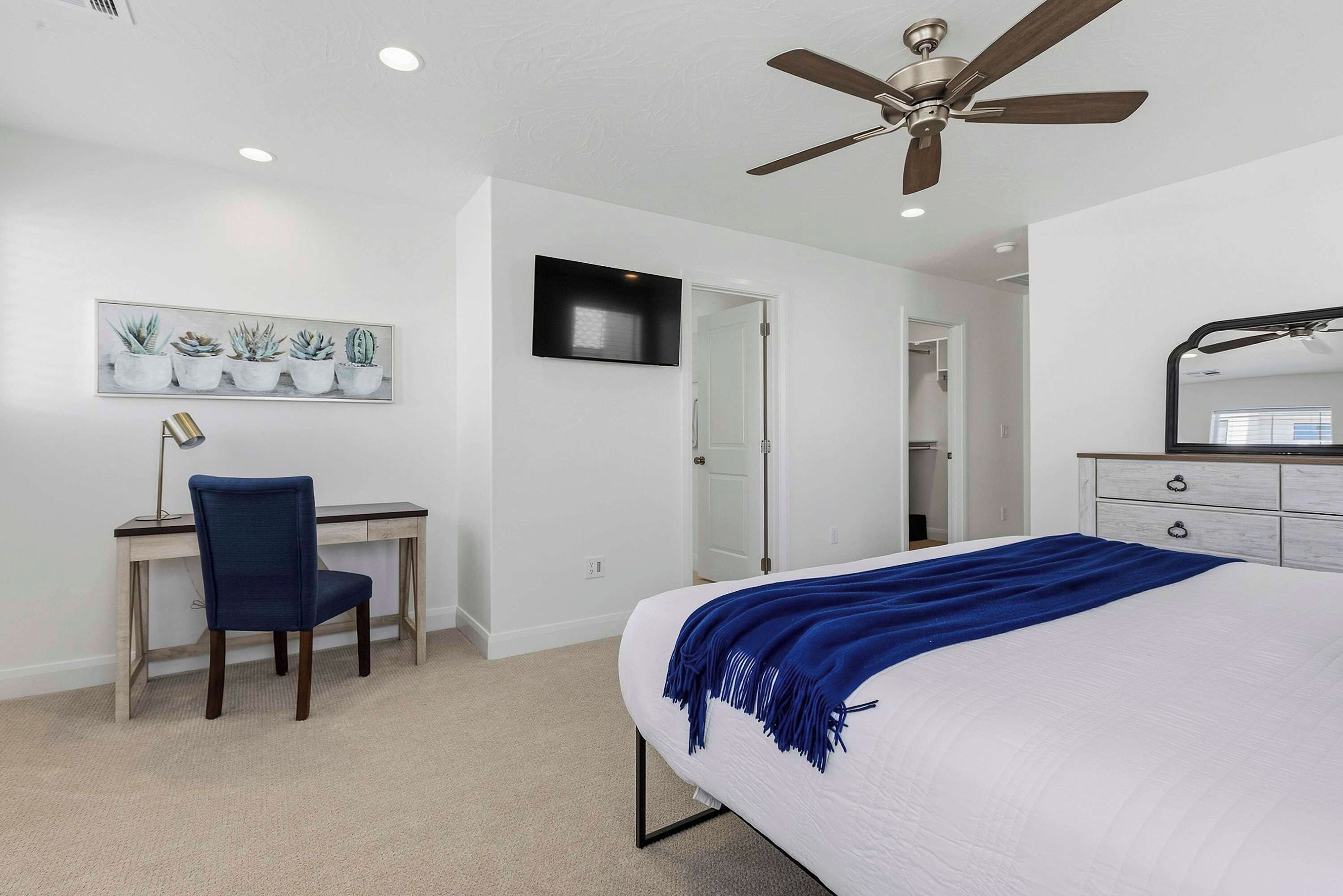 A bedroom with a bed, a desk, a chair, a ceiling fan, and a TV mounted on the wall.
