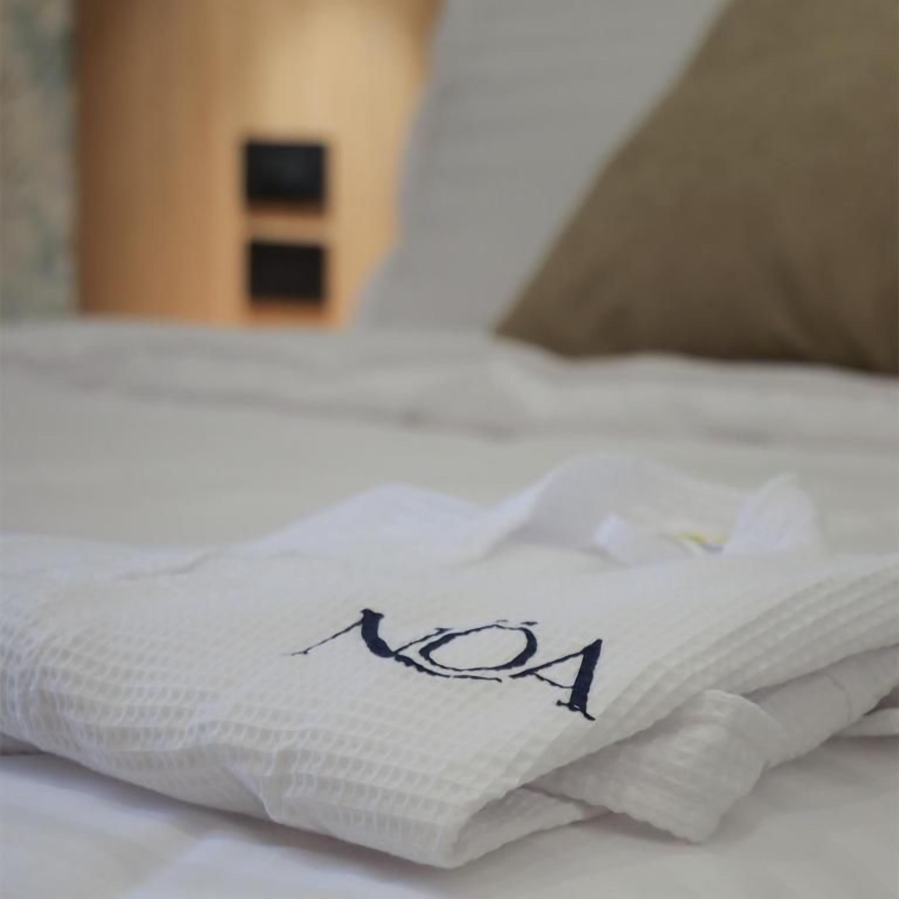 Una toalla blanca doblada con cuidado con el texto 'NOA', colocada sobre una cama con un cojín color beige al fondo.