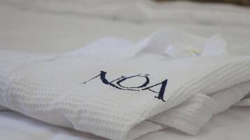 Una toalla blanca doblada con cuidado con el texto 'NOA', colocada sobre una cama con un cojín color beige al fondo.
