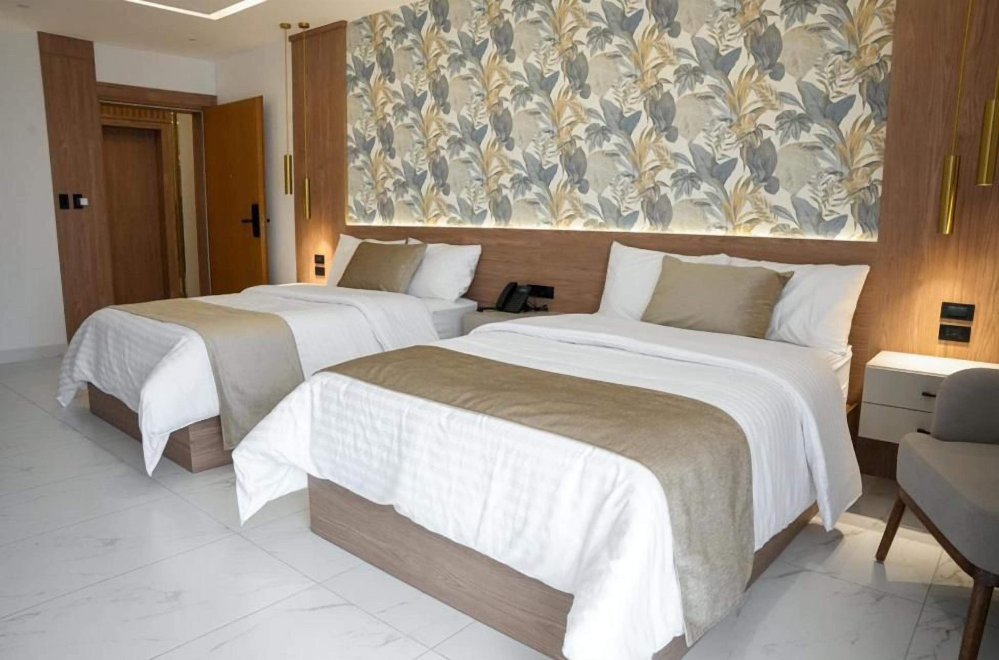 Quarto de hotel com duas camas, cabeceira de madeira, papel de parede floral e luminária moderna.