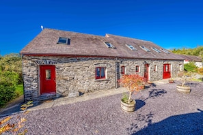 A stone cottage with a red door and multiple windows, surrounded by gravel and potted plants.