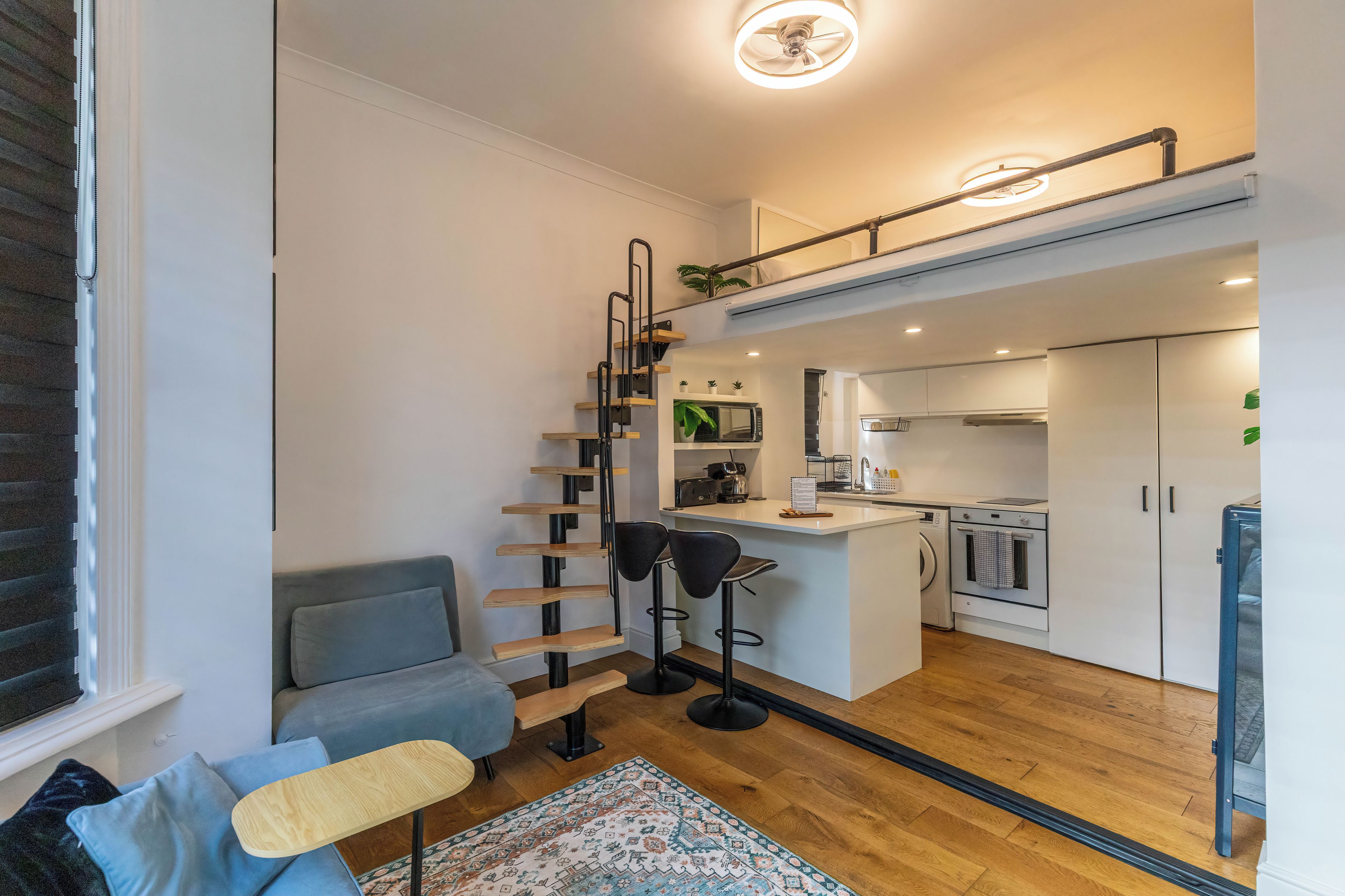 A compact living space with a kitchenette, a sofa, a small table, and a staircase leading to an upper level.