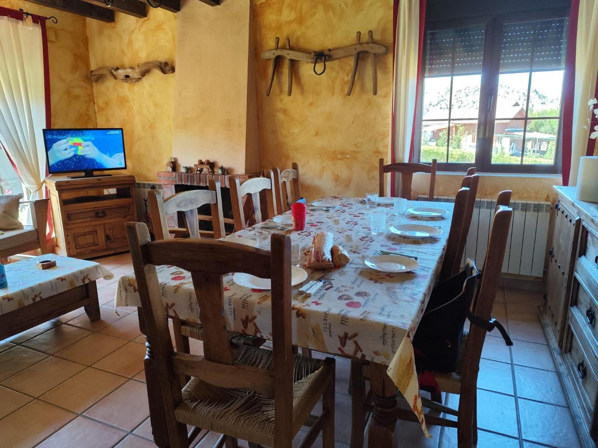 A dining area with a table set for four, wooden chairs, a television on a wooden cabinet, and a window with blinds.