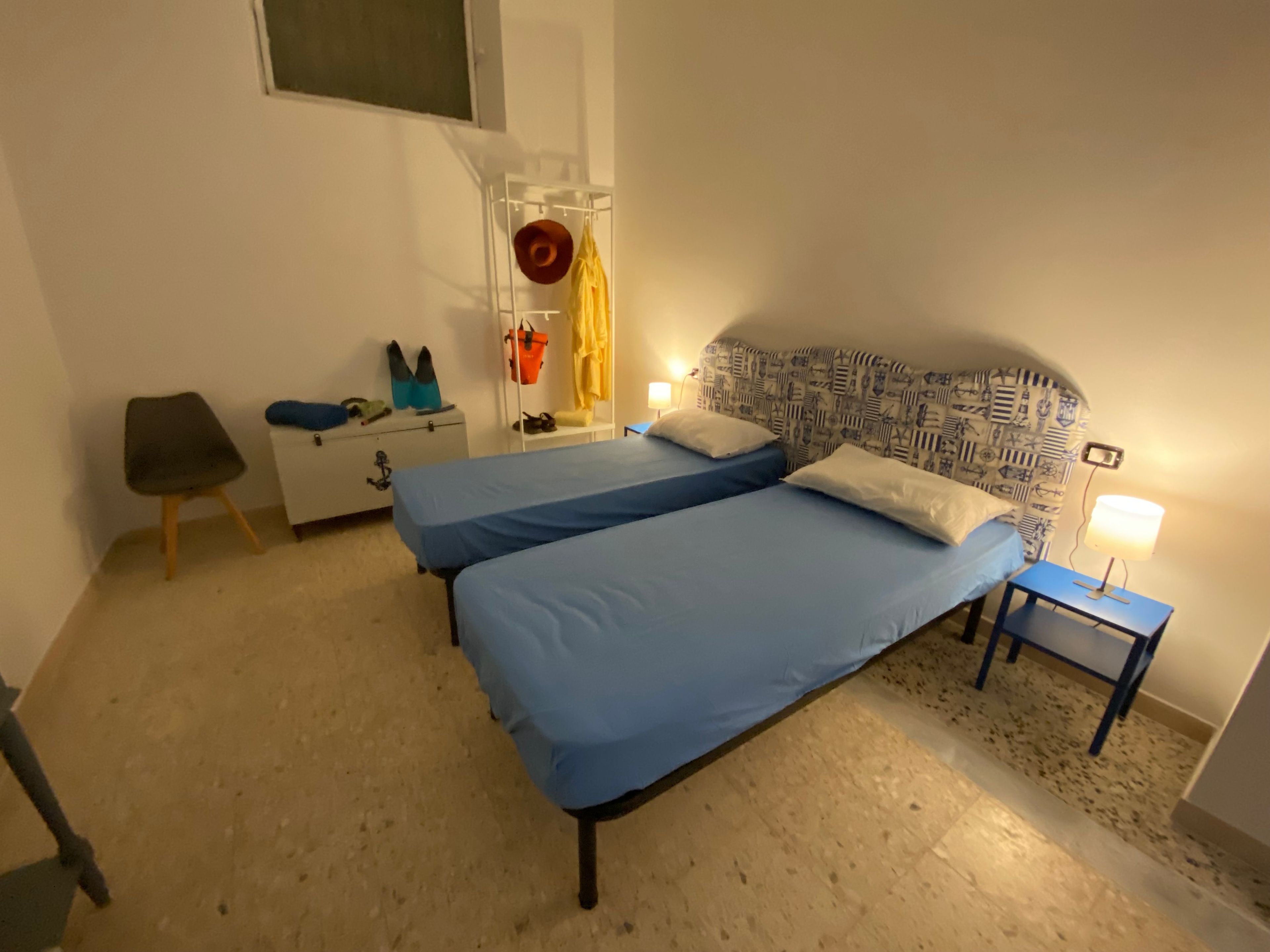 Et soveværelse med to senge, et lille bord med lampe, en stol, et garderobeskab og en reol med personlige ejendele.
