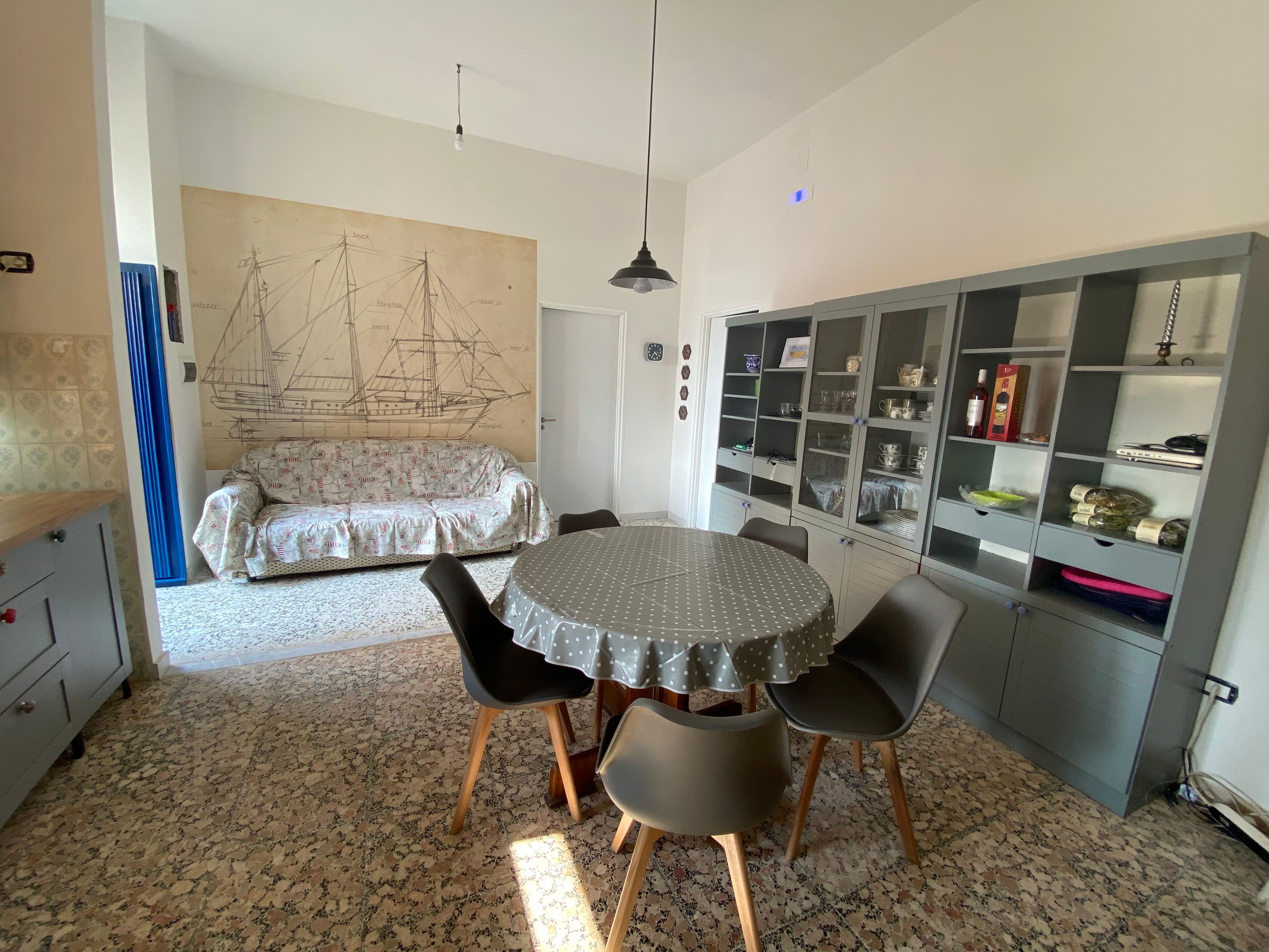 A dining area with a round table, chairs, a patterned sofa, and a wall with a ship illustration.