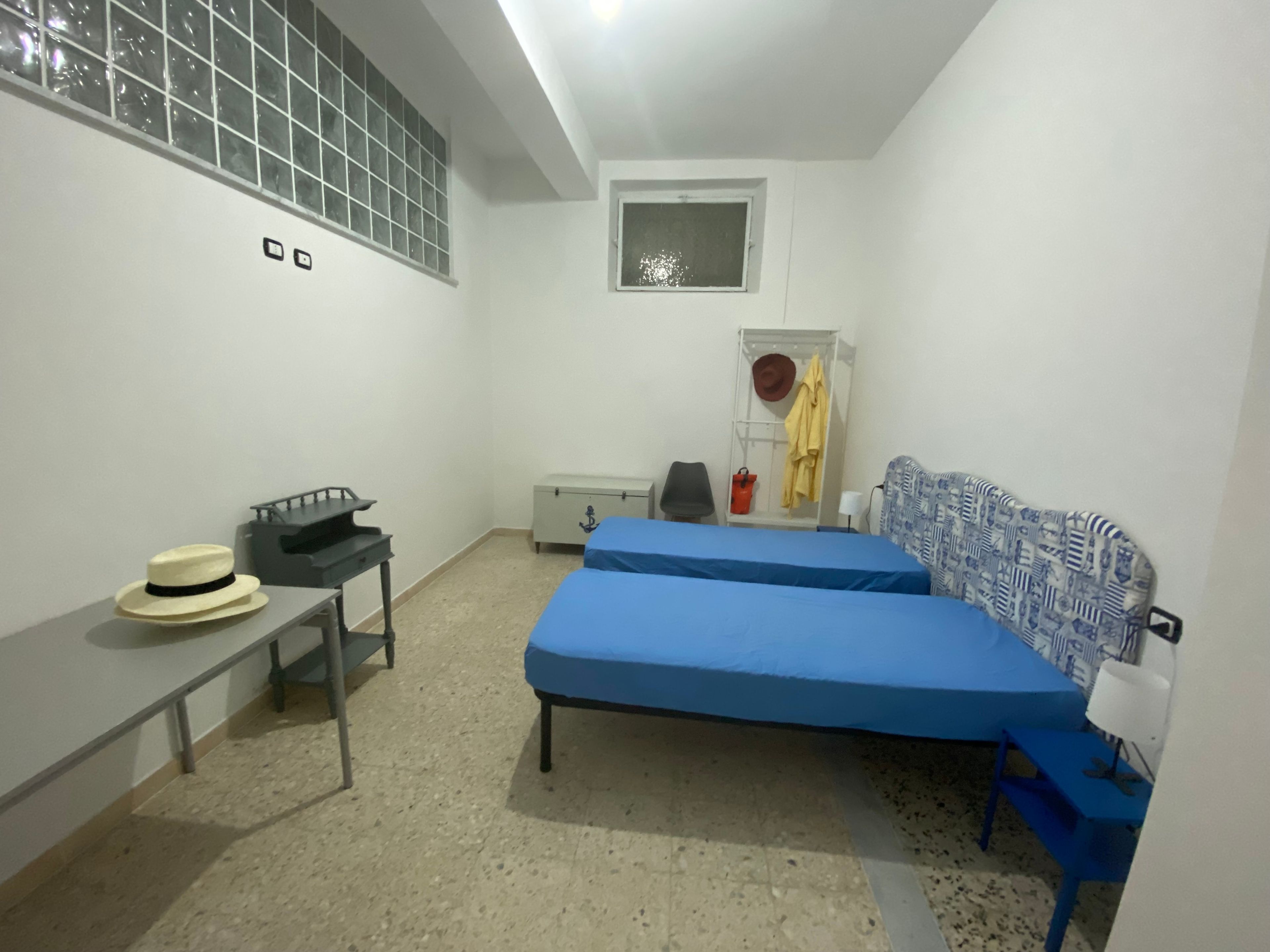 Quarto pequeno e simples com duas camas, uma escrivaninha, uma cadeira e uma janela.