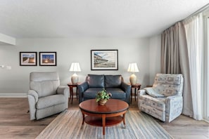Ein Wohnzimmer mit grauem Sofa, blauem Sofa, einem gemusterten Sessel, einem runden Couchtisch und zwei Lampen. An der Wand hängen gerahmte Bilder.