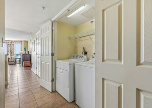 A laundry room with a washing machine and dryer, a tiled floor, and a door leading to a hallway. - Malibu Pointe 1001 -I/O Pools-Lazy River-4BR/4BA (North Myrtle Beach)