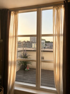 Una habitación con un gran ventanal, cortinas, un balcón pequeño con una planta y un coche de juguete.