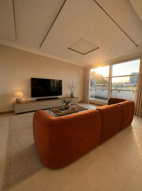 Una sala de estar moderna con un sofá grande color naranja, un televisor de pantalla plana, una mesa de centro y vistas de la ciudad a través de amplios ventanales.