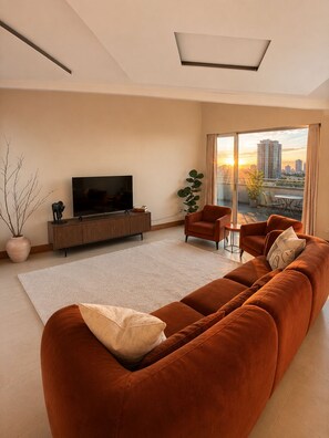 Ein modernes Wohnzimmer mit einer großen Couch, einem Fernseher und Blick auf eine Stadtkulisse durch ein großes Fenster.