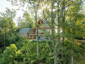 Eine Hütte, von dichtem Wald umgeben, mit einer markanten Holzkonstruktion und einem großen Fenster mit Blick auf das Grün.