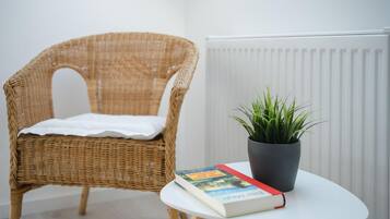 Een rieten stoel met een witte kussen, een bloempot en een boek op een klein wit tafeltje.
