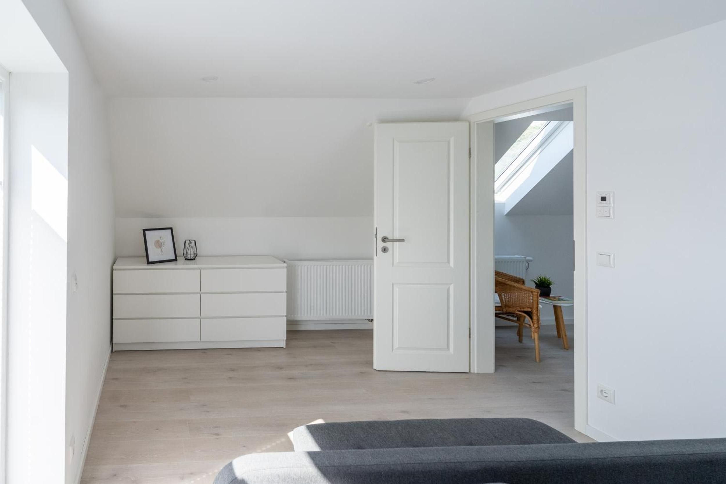 Una habitación amplia con techo alto, un armario blanco, un radiador y una puerta que da a otra estancia con una mesa y una silla.
