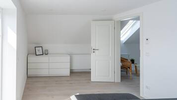 Una habitación amplia con techo alto, un armario blanco, un radiador y una puerta que da a otra estancia con una mesa y una silla.