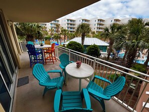 Ein Balkon mit türkisfarbenen Stühlen und einem kleinen Tisch, mit Blick auf einen Poolbereich mit Palmen und einem Gebäude im Hintergrund.