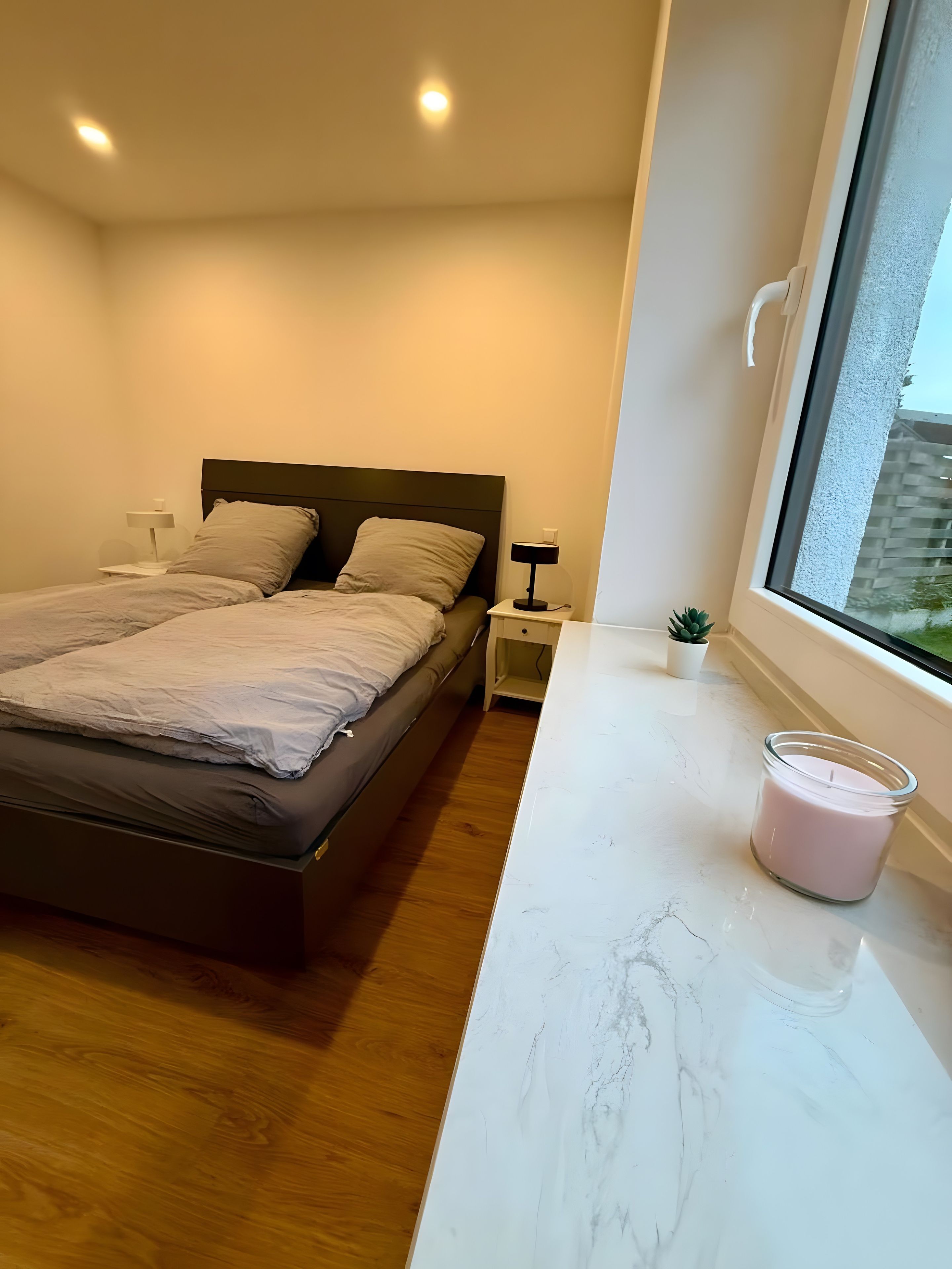 A bedroom with a bed, bedside tables, a nightstand, a window, and a small plant.