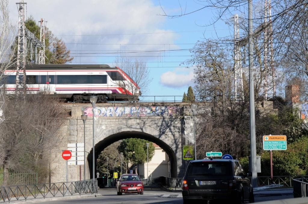 Un tren pasando debajo de un puente con grafitis, el rótulo de una farmacia y una señal de prohibido el paso.