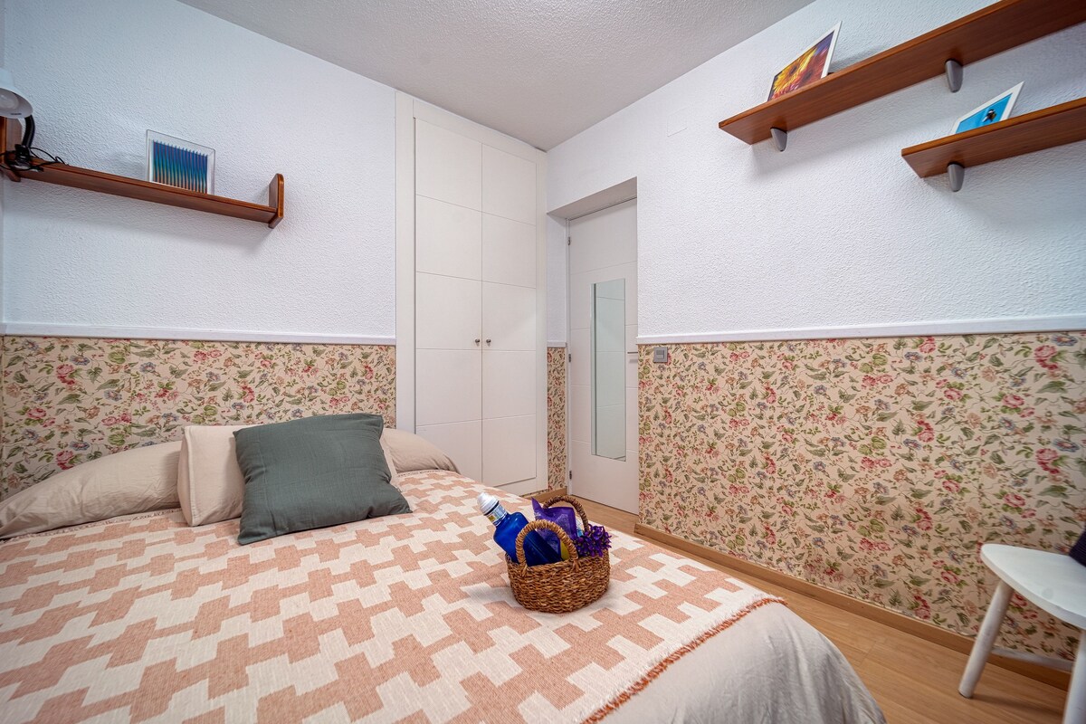 A bedroom with a bed, pillows, a bedside table, a wall-mounted shelf, and a door.