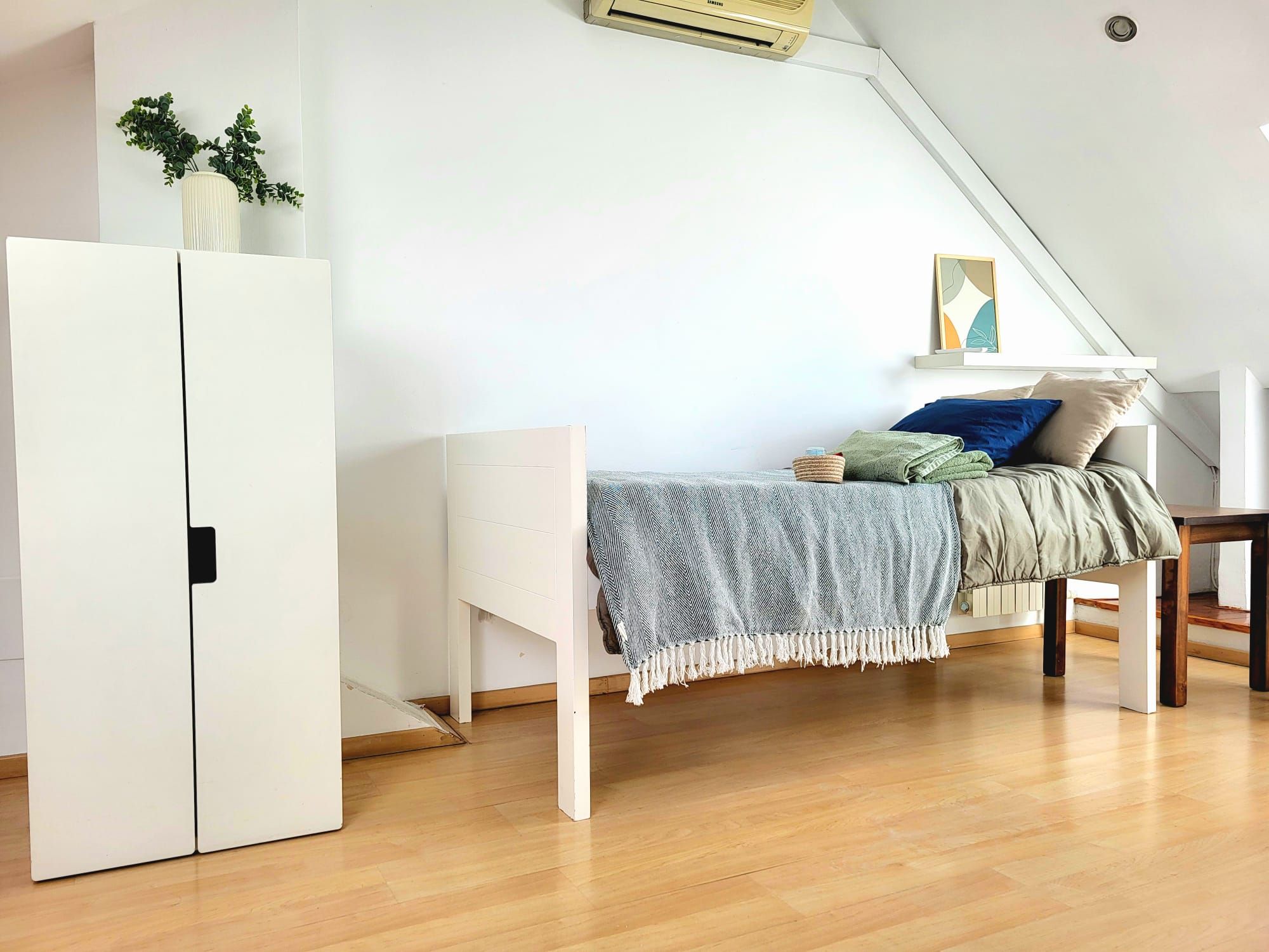 Una habitación pequeña y bien iluminada con una cama, un armario blanco y una planta en la pared.