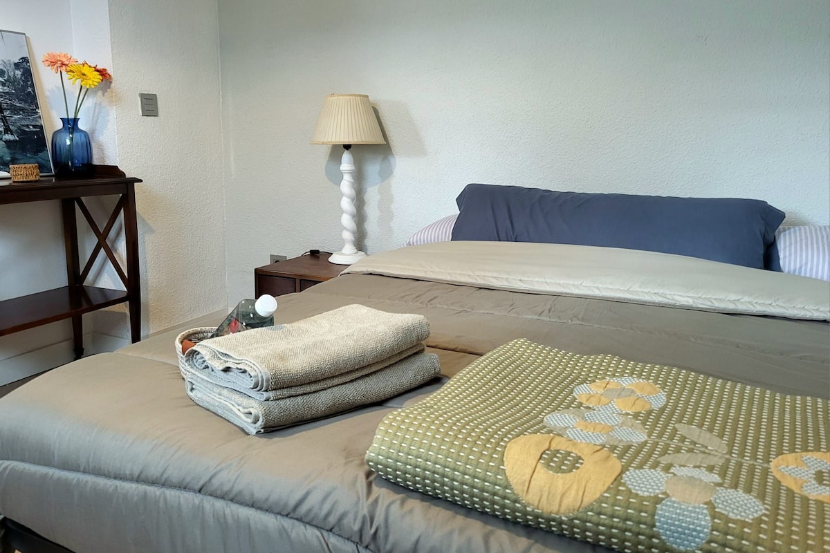 A neatly made bed with pillows and folded towels, a bedside table with a lamp, and a wall-mounted power outlet.