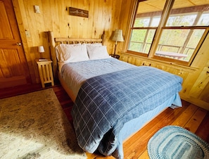A wooden cabin bedroom with a bed, nightstand, lamp, and a window with a view of trees.