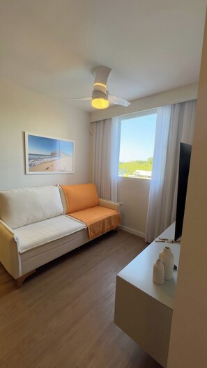 A hotel room with a white sofa, a ceiling fan, a painting of a beach, a large window with curtains, and a flat-screen TV on a white table. - Cozy apartment in Barra Olímpica, close to the beaches. (Rio de Janeiro)