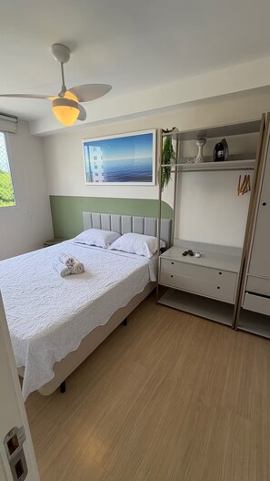 A bedroom with a bed, a ceiling fan, a painting, a wardrobe, and a small table. - Cozy apartment in Barra Olímpica, close to the beaches. (Rio de Janeiro)