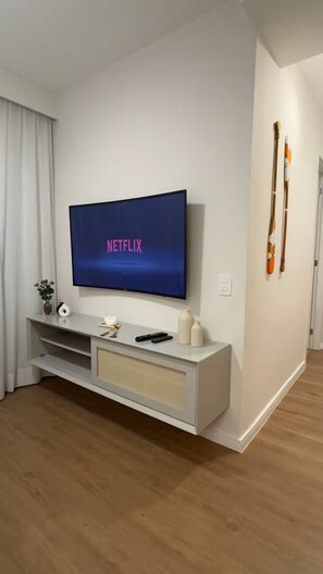A modern living room with a flat-screen TV displaying 'NETFLIX' on the screen, a white entertainment unit with a remote, and a door with a wooden door knocker. - Cozy apartment in Barra Olímpica, close to the beaches. (Rio de Janeiro)