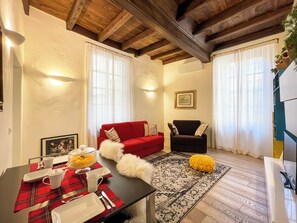 Ein gemütliches Wohnzimmer mit einem roten Sofa, einem schwarzen Sessel, einem Esstisch mit rot-weiß kariertem Tischläufer und einem gemusterten Teppich.