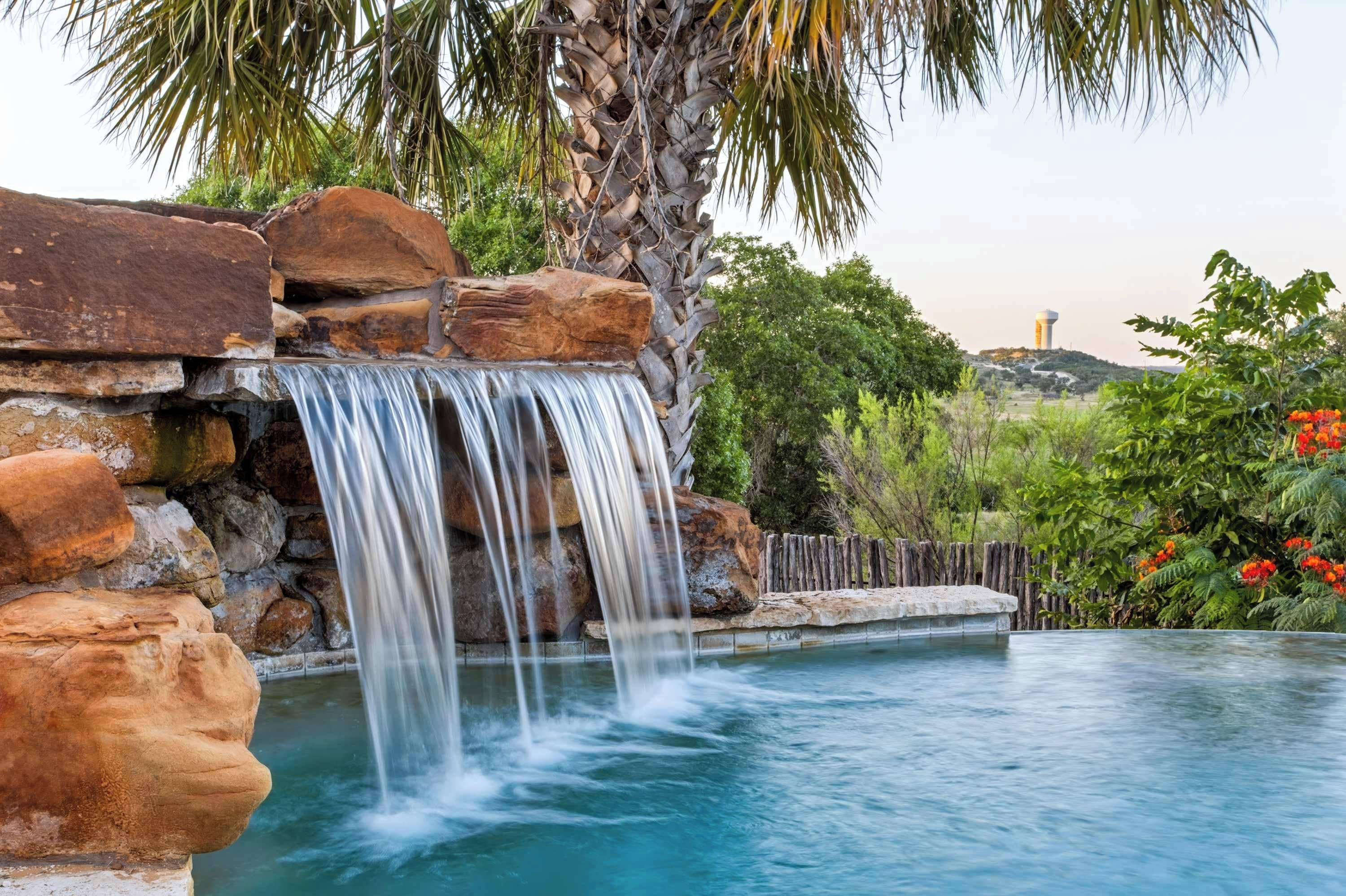 Formación rocosa natural con una cascada que cae en una piscina cristalina, rodeada de exuberante vegetación y una torre de agua al fondo.