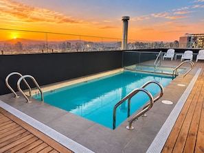 A rooftop pool with clear blue water, surrounded by wooden deck and metal railings, with a city skyline in the background. - AC - Estadio Nacional - Estacionamiento (Ñuñoa)