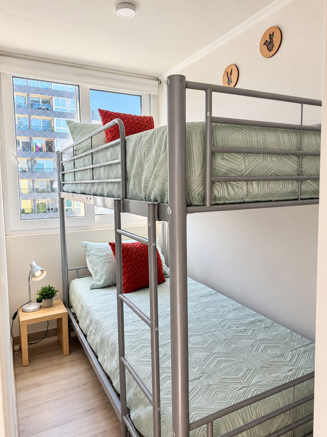 A bunk bed with a geometric patterned mattress, a bedside table with a lamp, and a window with a view of a building.