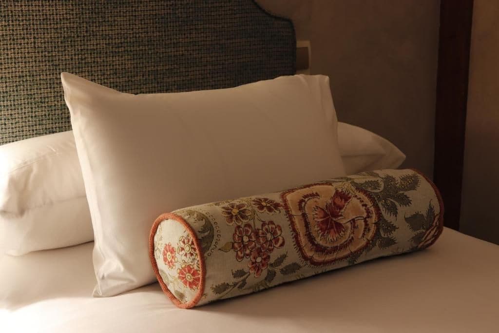 Een bed met een wit kussen en een opgerold decoratief kussen met bloemenmotieven.