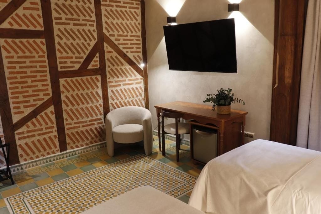 Una habitación de hotel con pared de ladrillo estampada, un televisor de pantalla plana montado sobre un escritorio de madera, una silla y una cama con cubrecama blanco.