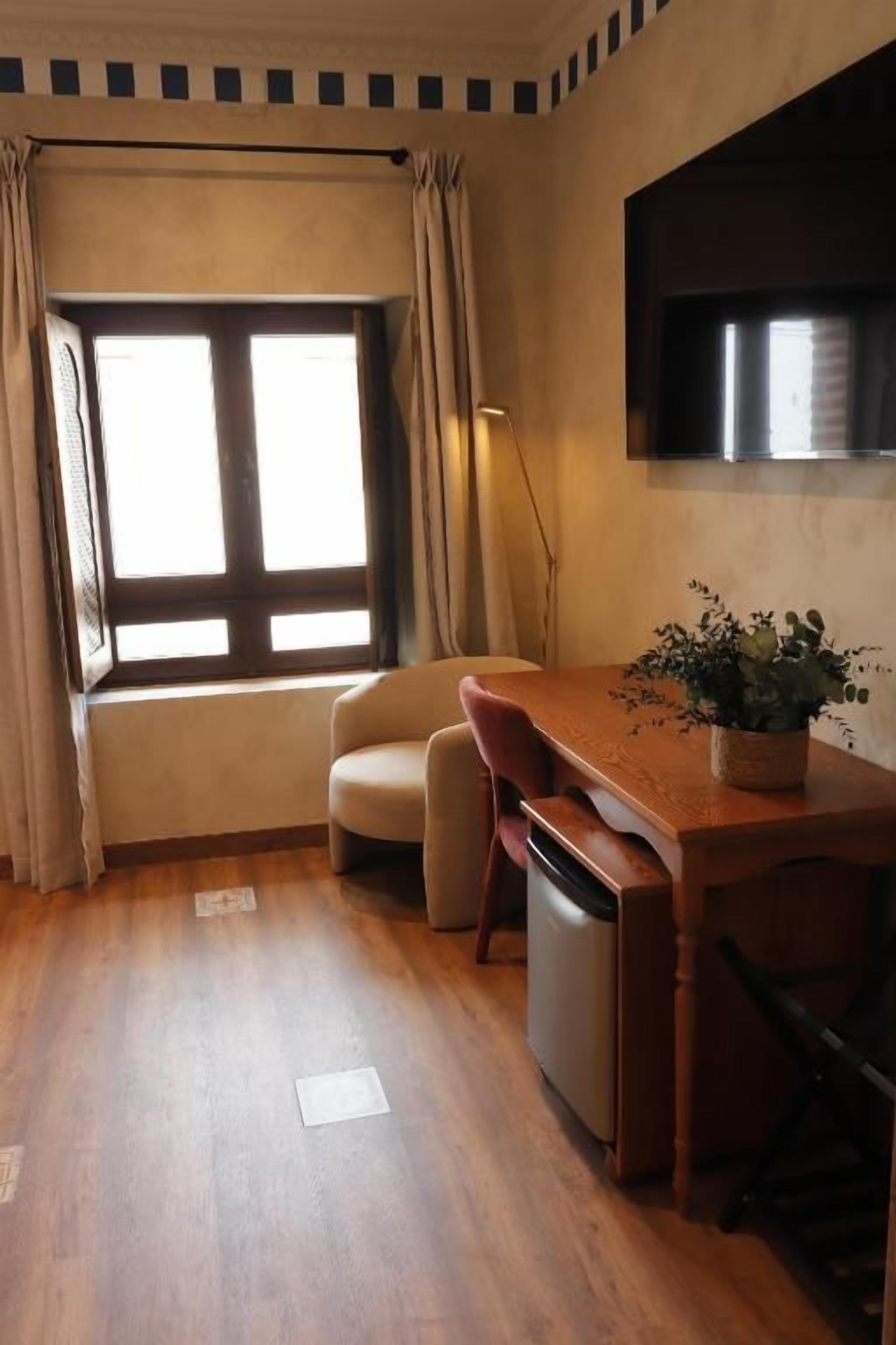 A compact room with a wooden floor, a small sofa, a dining table with a potted plant, a refrigerator, and a window with curtains.