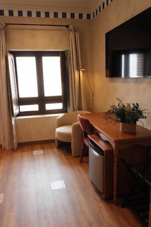 A compact room with a wooden floor, a small sofa, a dining table with a potted plant, a refrigerator, and a window with curtains.